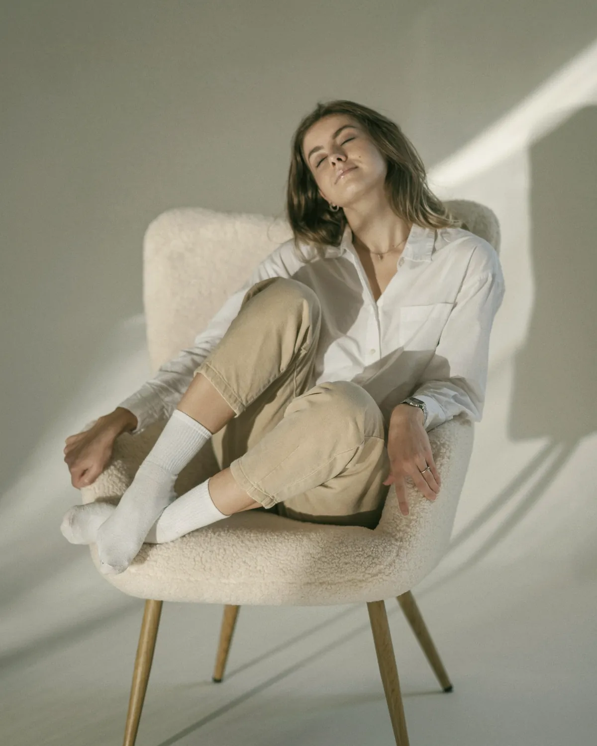 Mujer relajada en un sillón blanco, con camisa blanca y pantalón beige